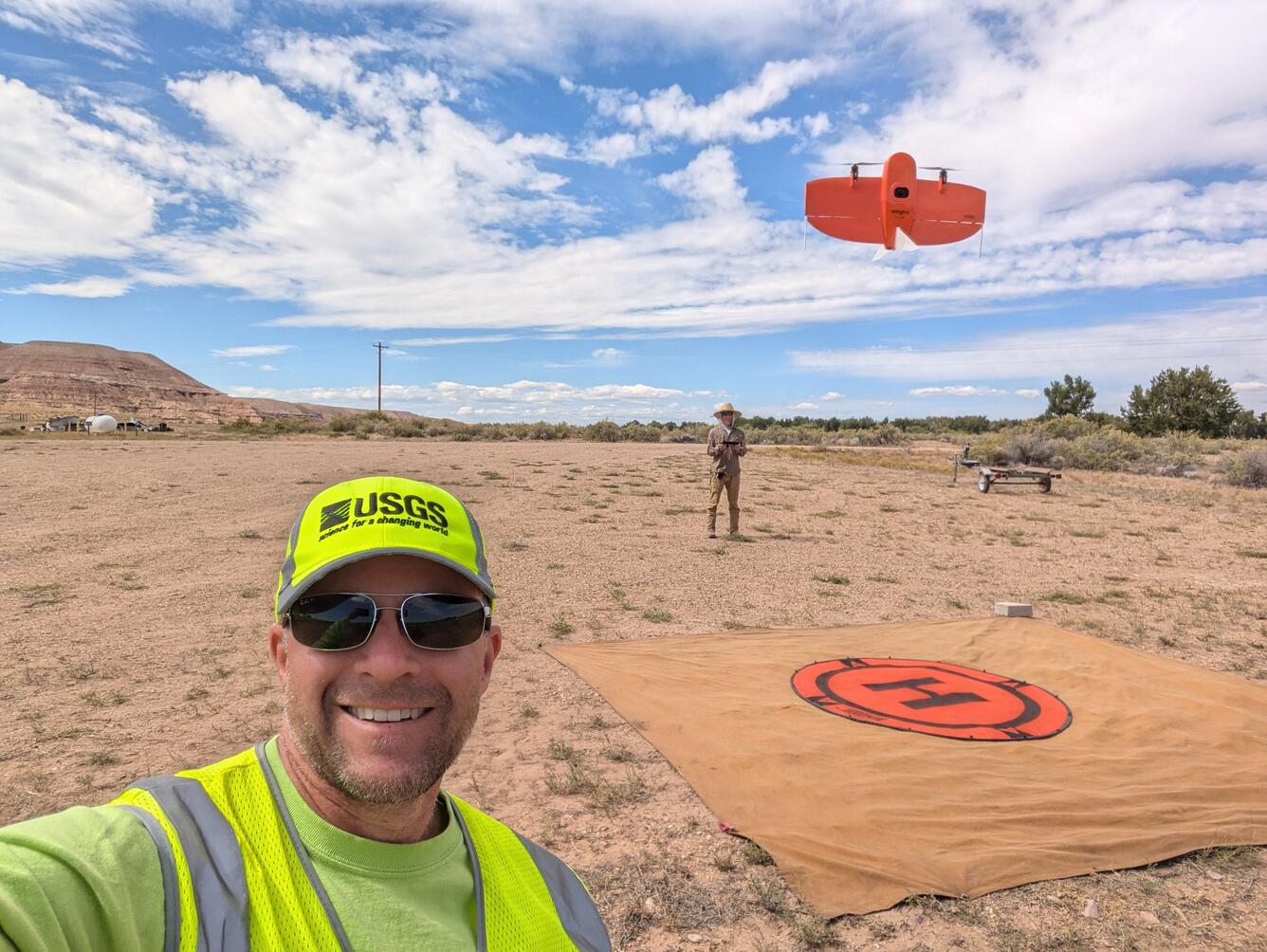 two people stand in a dirt landscape with a UAS in the air and helipad on the ground
