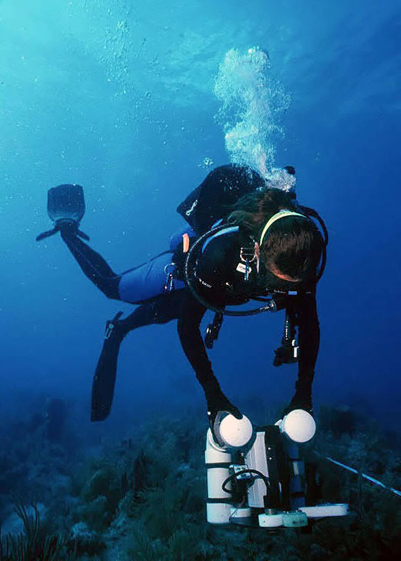 A woman dives in dark blue water holding a white machine below her, near a coral reef.
