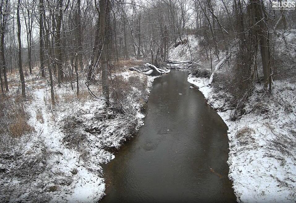 Winter snow blankets the grey and brown underbrush which frame the dark waters of the river flowing in from the distance