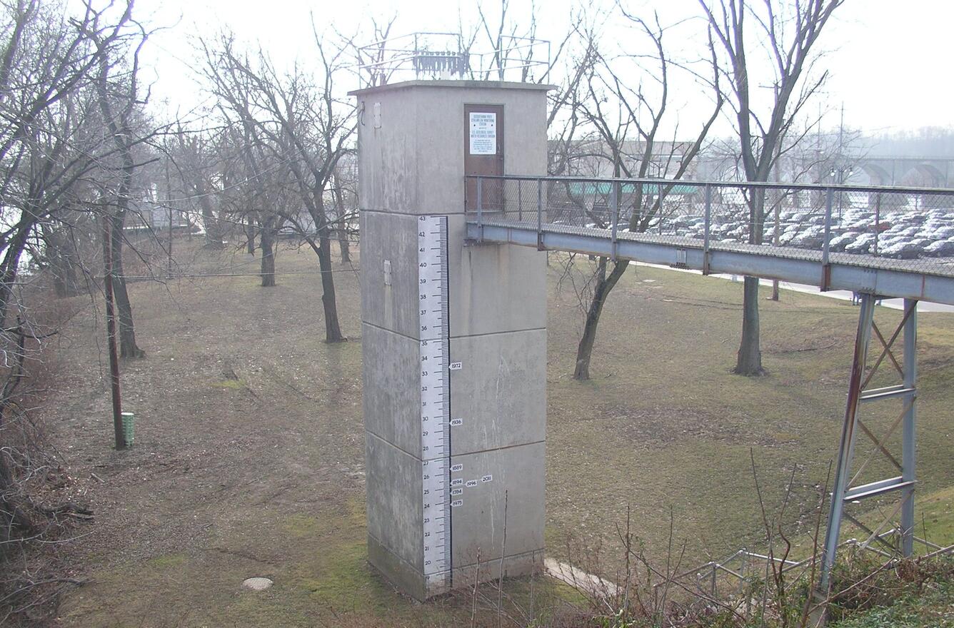 Photograph of stream floodplain with tall, narrow concrete building with gangplank from bank, ruler and markings on the side.