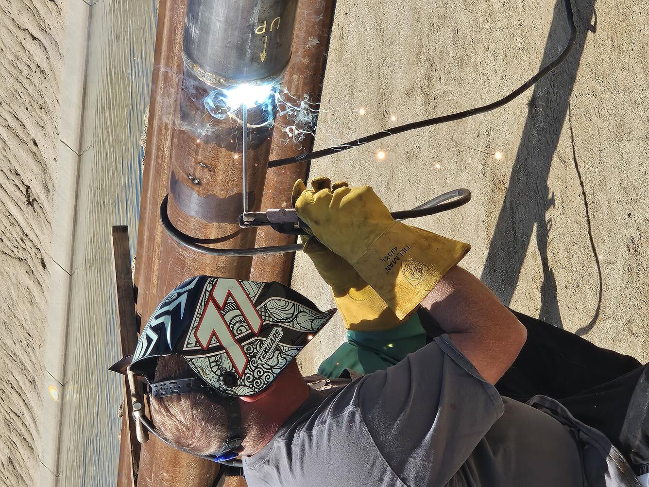 A welder in a helmet and heavy gloves welds a joint between two sections of steel well casing pipe on the ground beside a can