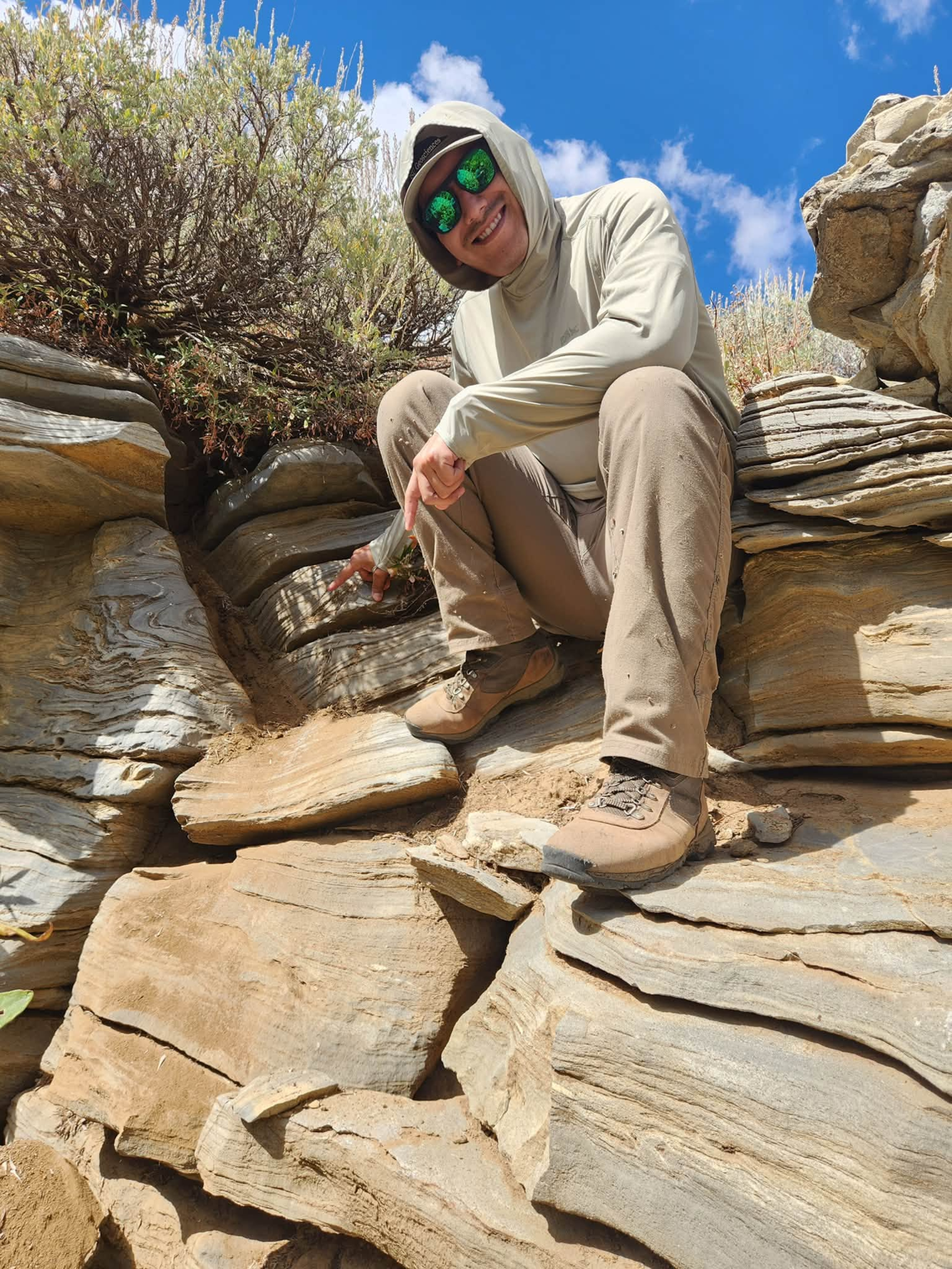 A person sits on top of an outcrop of brown rocks with bedding lines. 