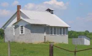 Image from the Amish America website of a simple Amish schoolhouse with white roof, red brick chimney, and an outhouse.