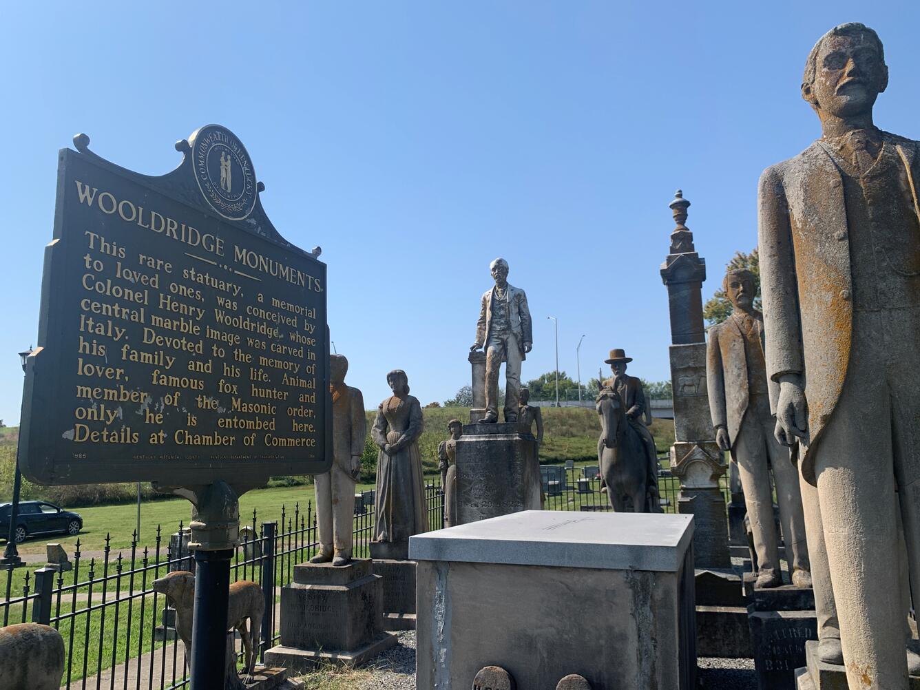 Historical marker installed beside weathered human-figure statues, all enclosed by an iron fence.