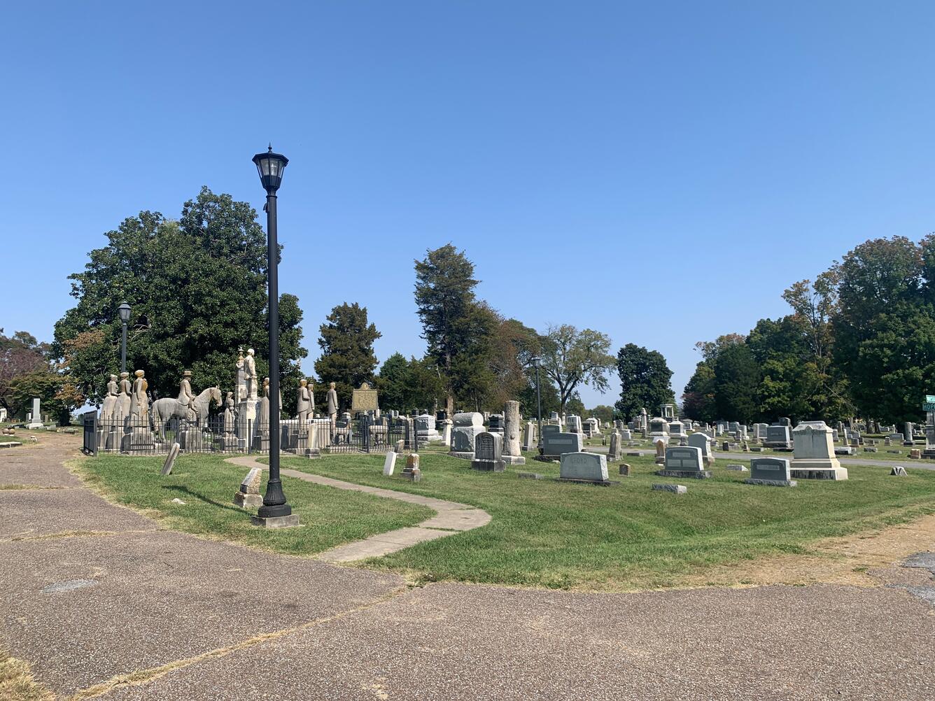A set of weathered statues visible from a cemetery road intersection, with a pathway leading up to the burial plot.