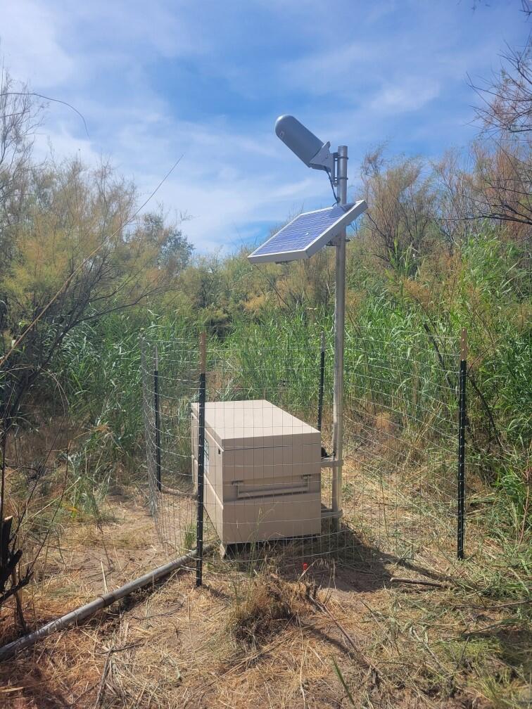 Metal chest, solar panel, and antenna set on grassy ground surrounded by wire fencing.