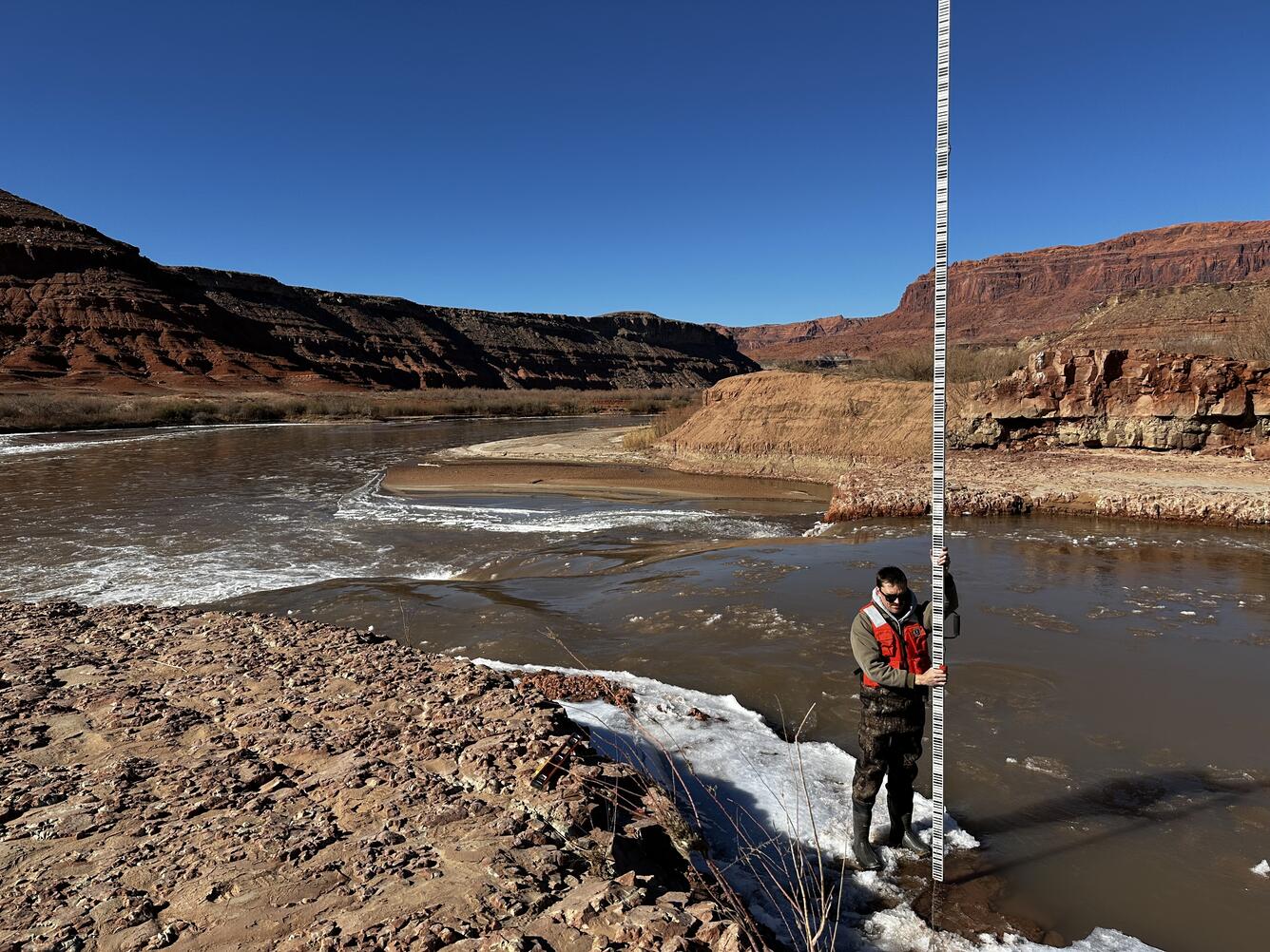 Sweeping view of wide meandering river in red geologic formations. Person in foreground holding very tall survey rod. 