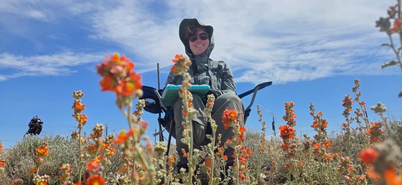 A person sitting in a chair with a clipboard smiles, there are orange globemallow flowers in the foreground and a cloudy blue sky in the background