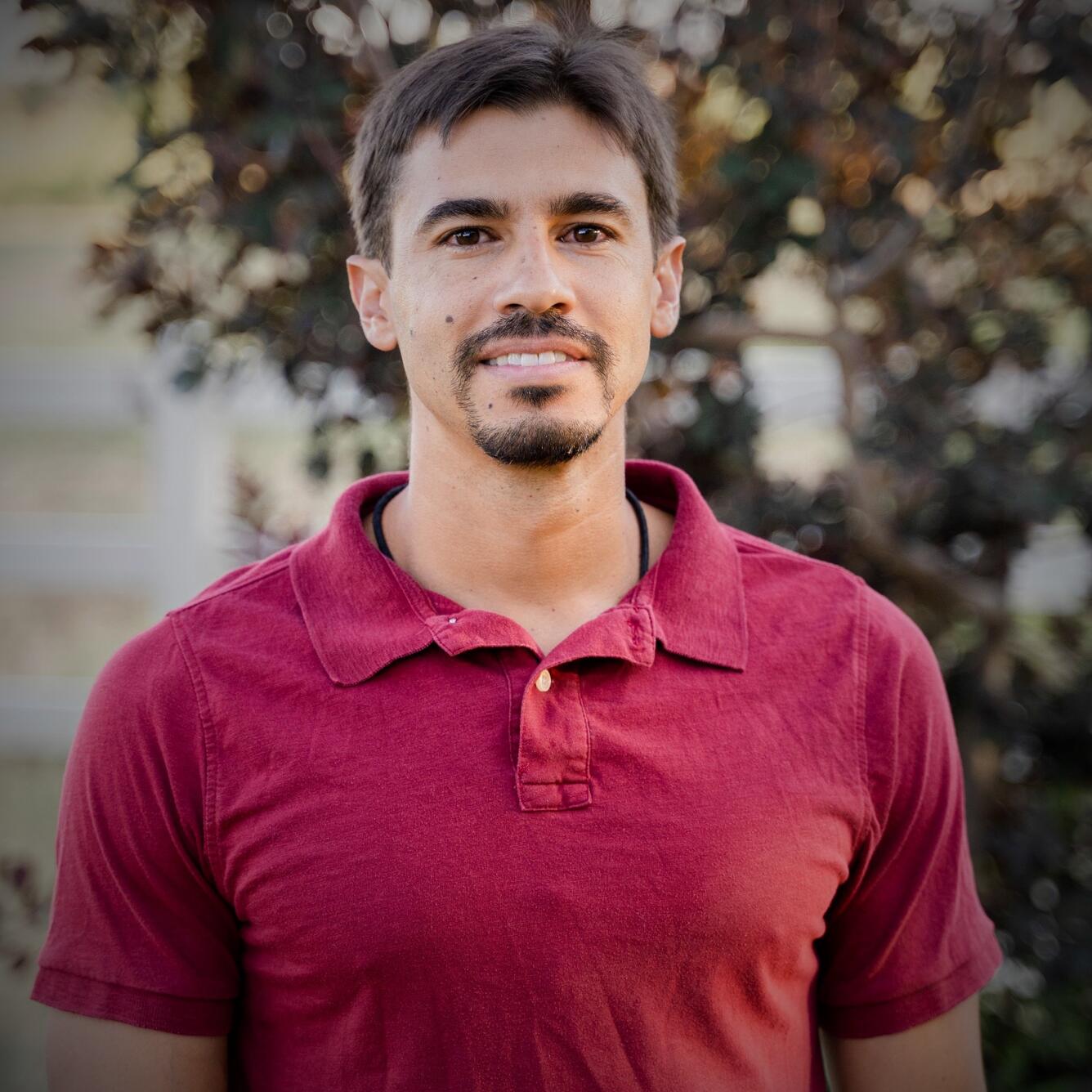 A man with short brown hair and a red polo shirt smiles at the camera. In the background, tree branches and foliage can be seen.