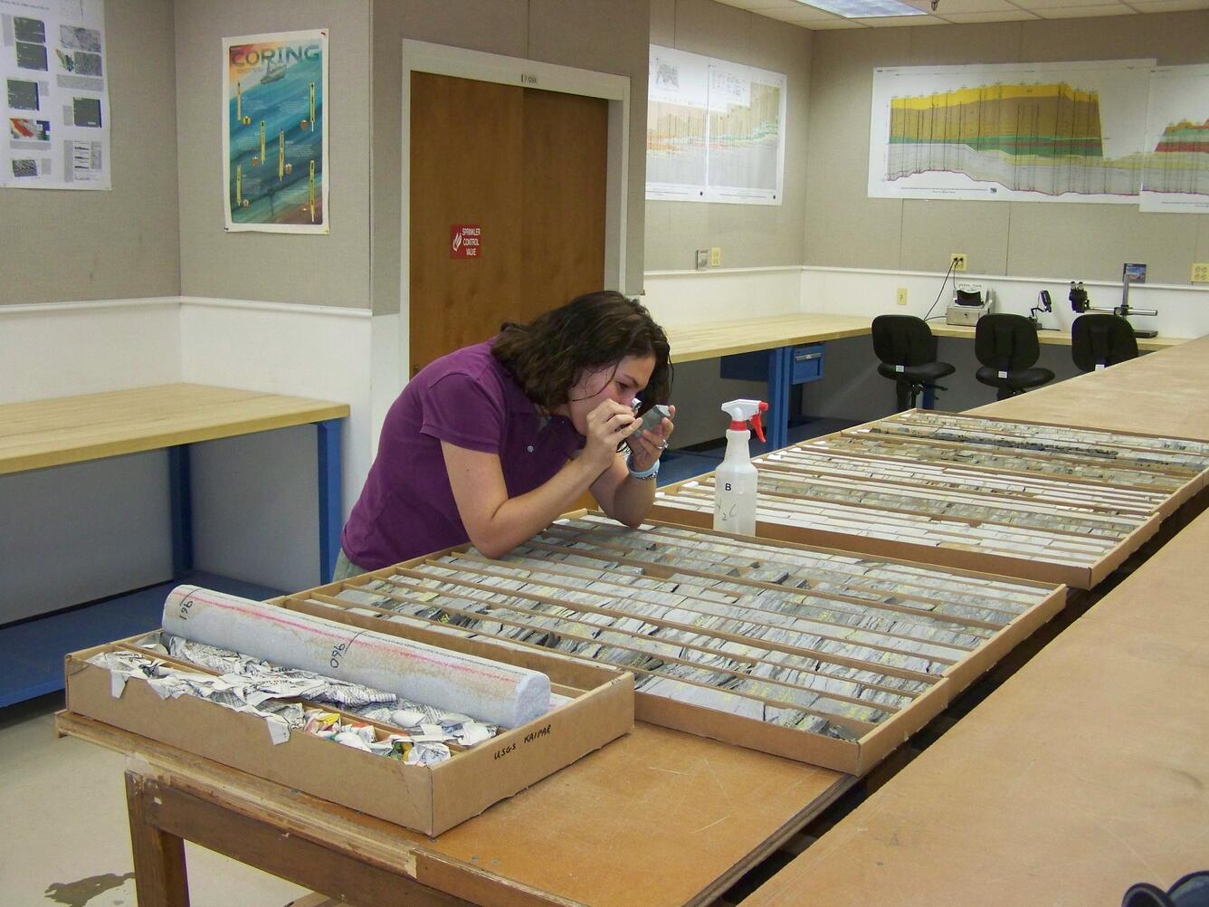 Woman in purple shirt looks at rock core through a hand lens