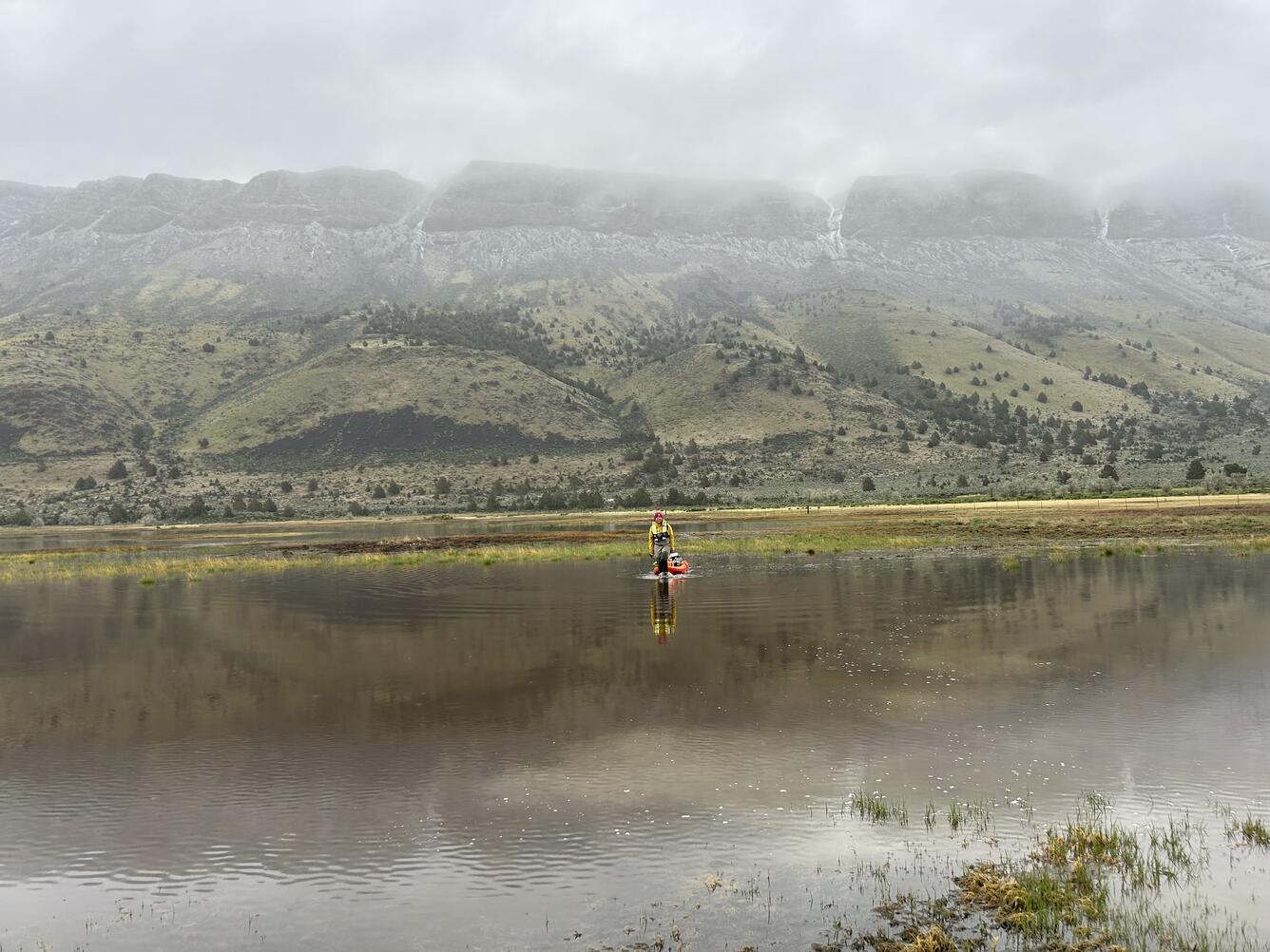 A female in safety gear pulling equipment through the water looking small amidst the stunning background of misty hills