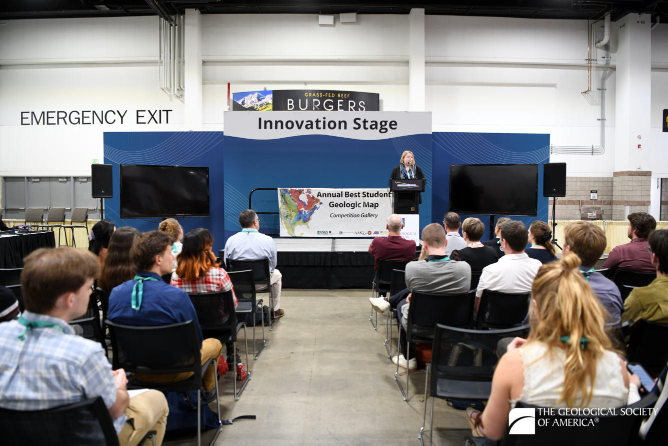 A person standing at a podium on a stage addresses a seated crowd. Behind presenter are two banners reading “Innovation Stage” and “Annual Best Student Geologic Map Competition Gallery.”