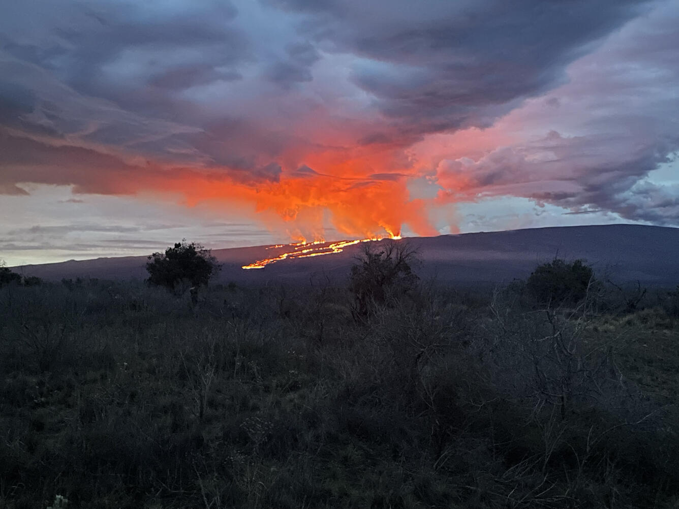 glowing red and orange lava flows moving down a dark slope with orange and red clouds in the sky.