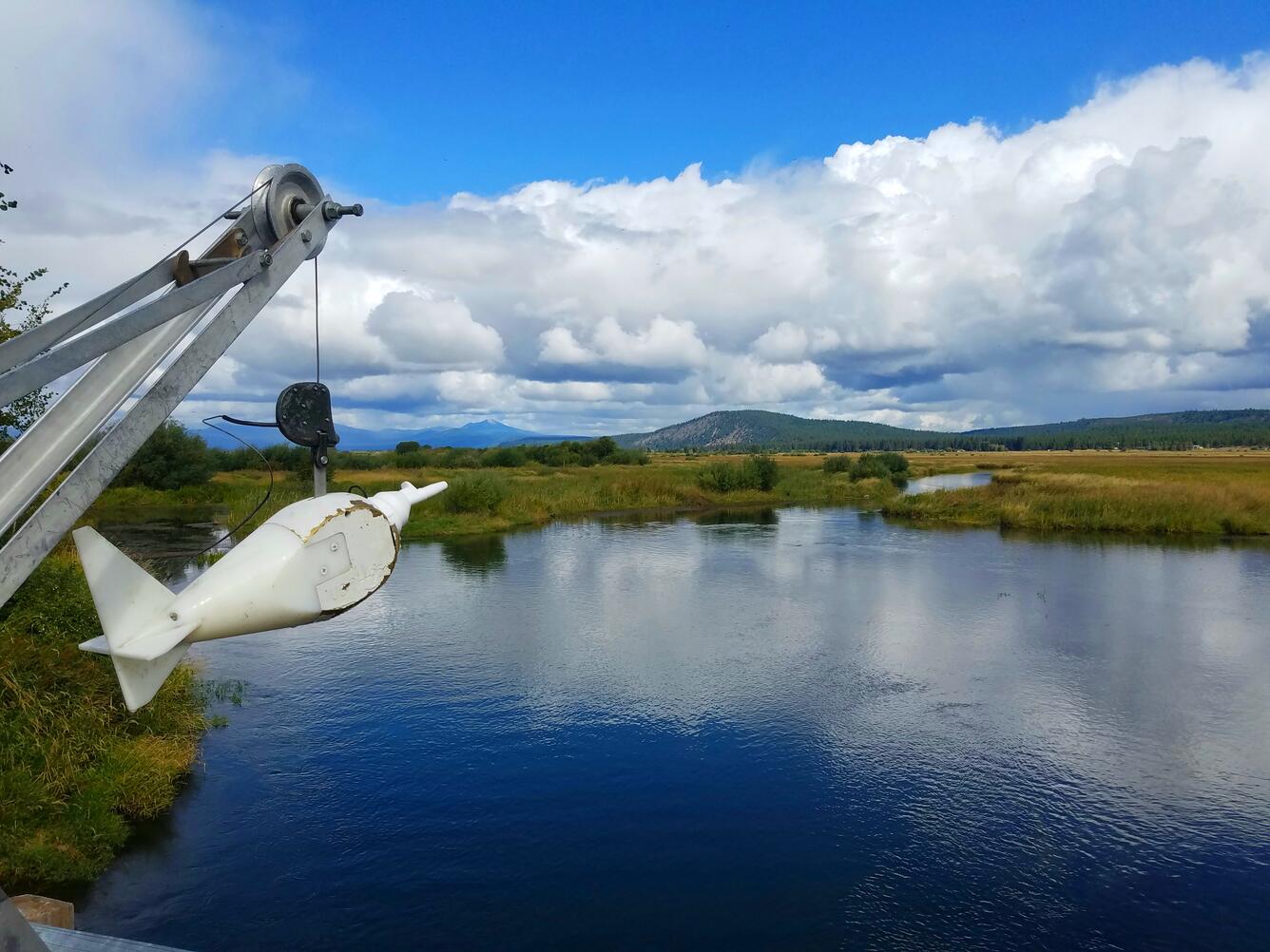 partly cloudy day. A white torpedo shaped sediment sampler hangs from a crane over a bridge with blue water below