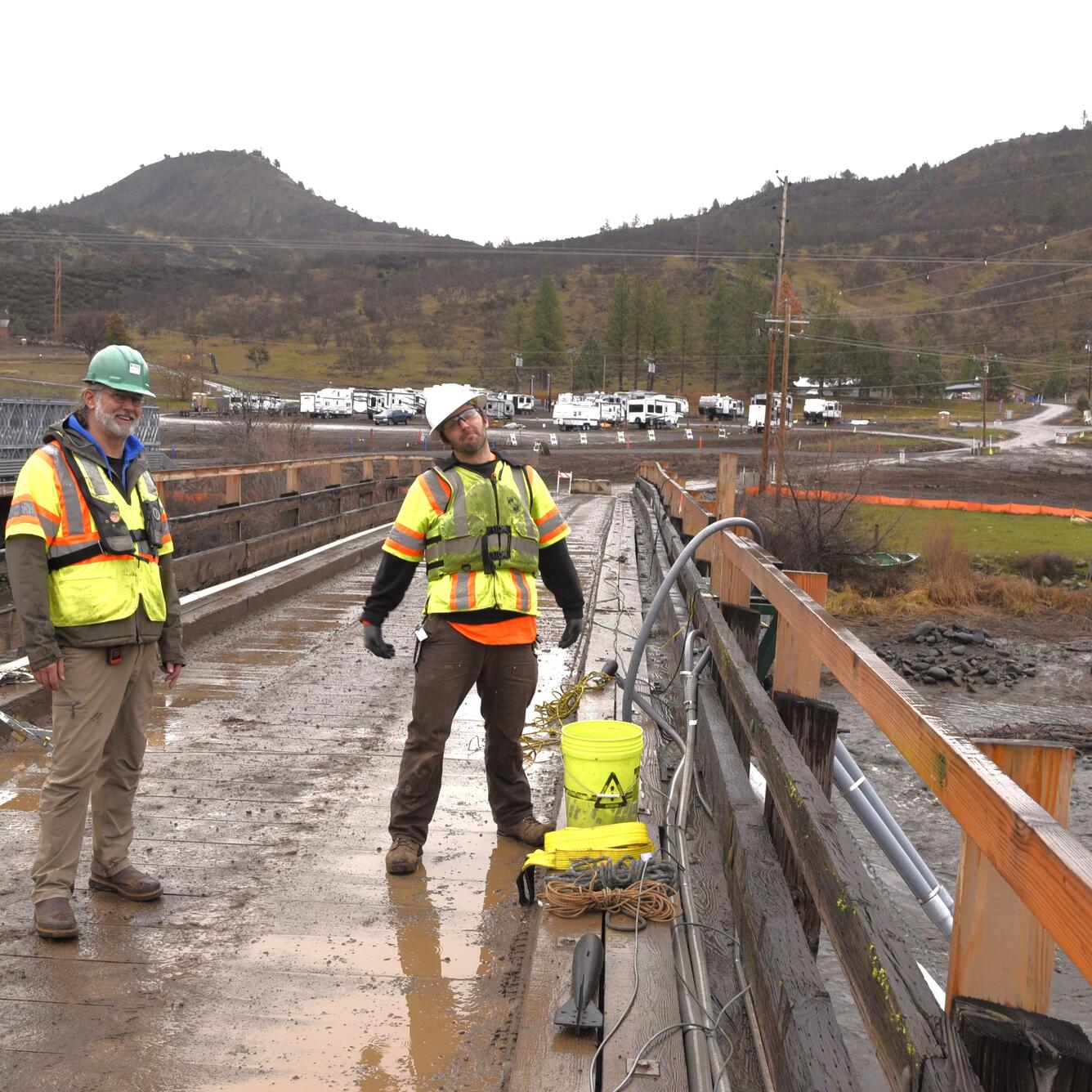 Two scientists in bright colored safety gear and hard hats stand on a bridge above the river.