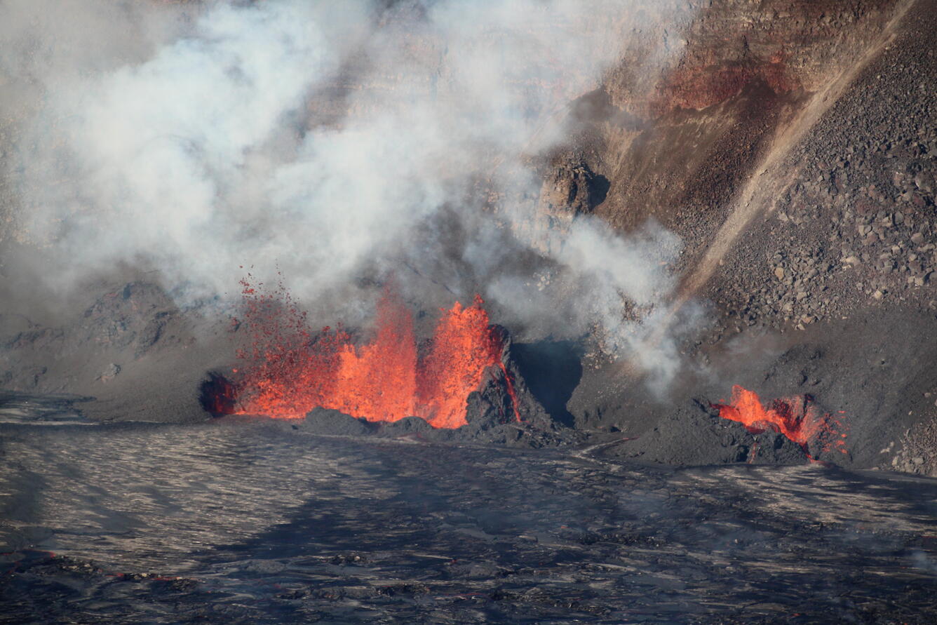 Orange lava fountains erupting inside of black lava spatter cone at the base of a crater wall