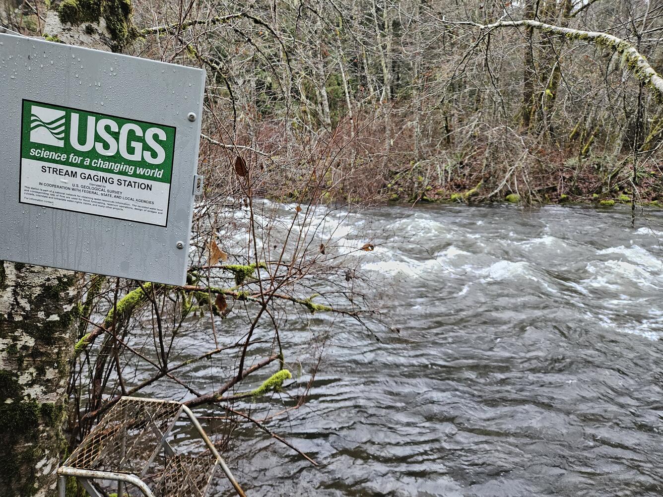 square metal enclosure door with USGS signage swings open by a rushing river