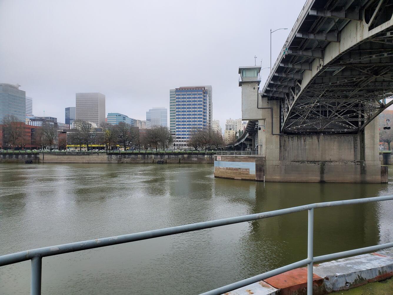 looking at SW Portland waterfront from middle of river. Brown water moves past bridge dolphin. Low clouds shroud skyscrapers