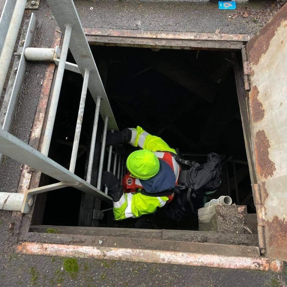 a person in bright safety gear descends a metal ladder through a square hole down into darkness