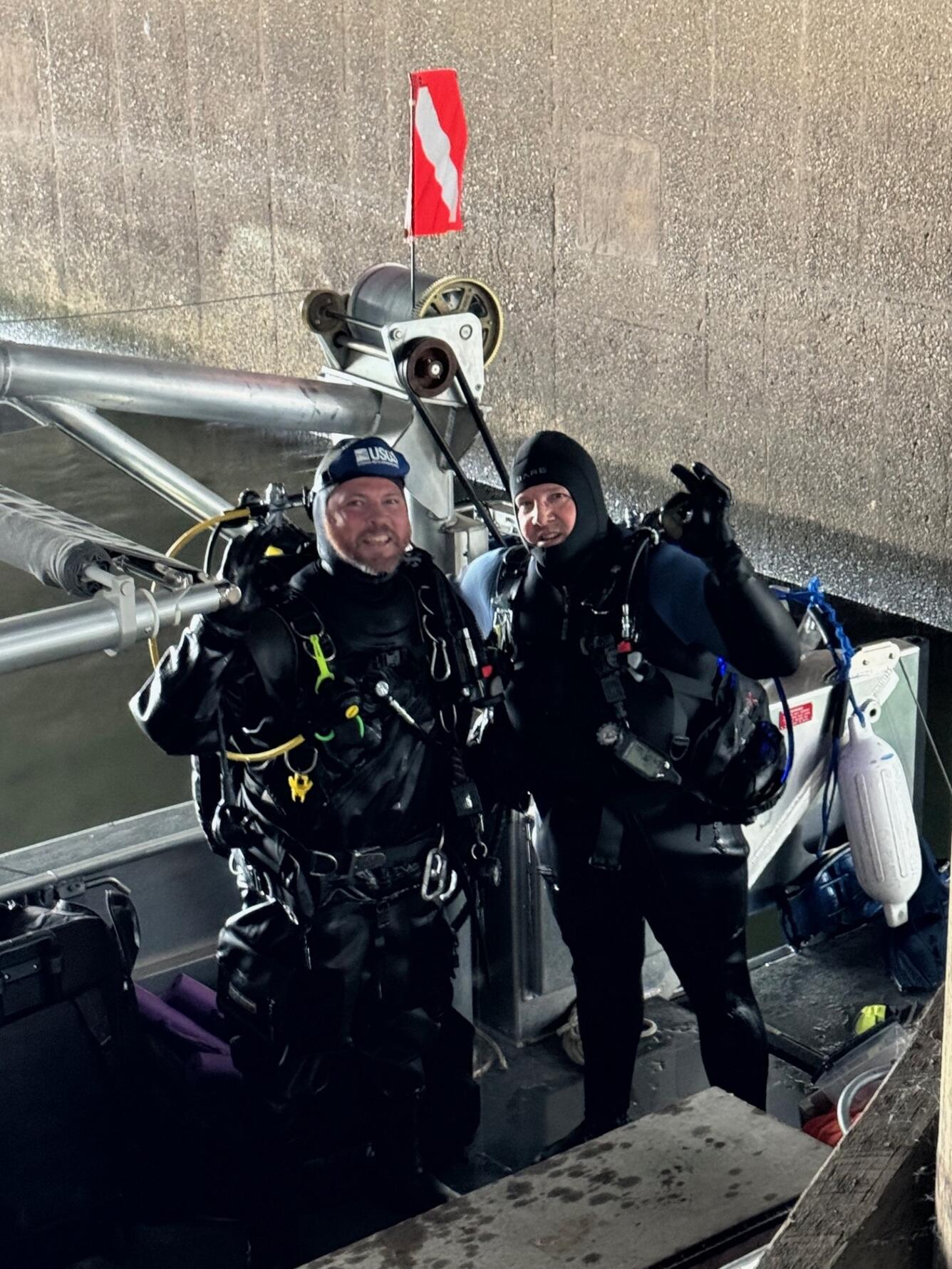 Two men in black wet suits with oxygen tanks smile and wave at the camera from a boat docked under a bridge.