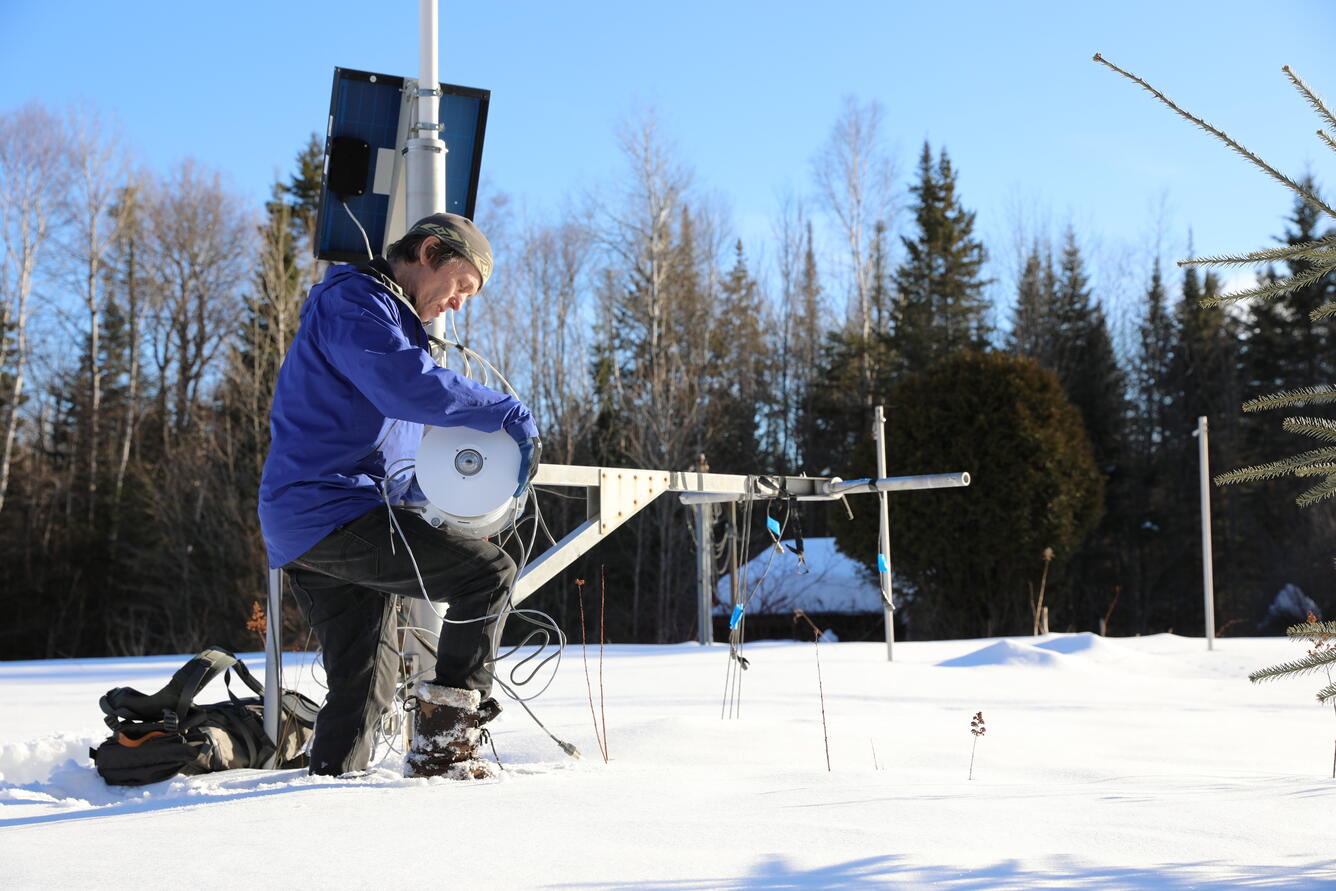 A man kneels down in the snow fixing metal equipment.