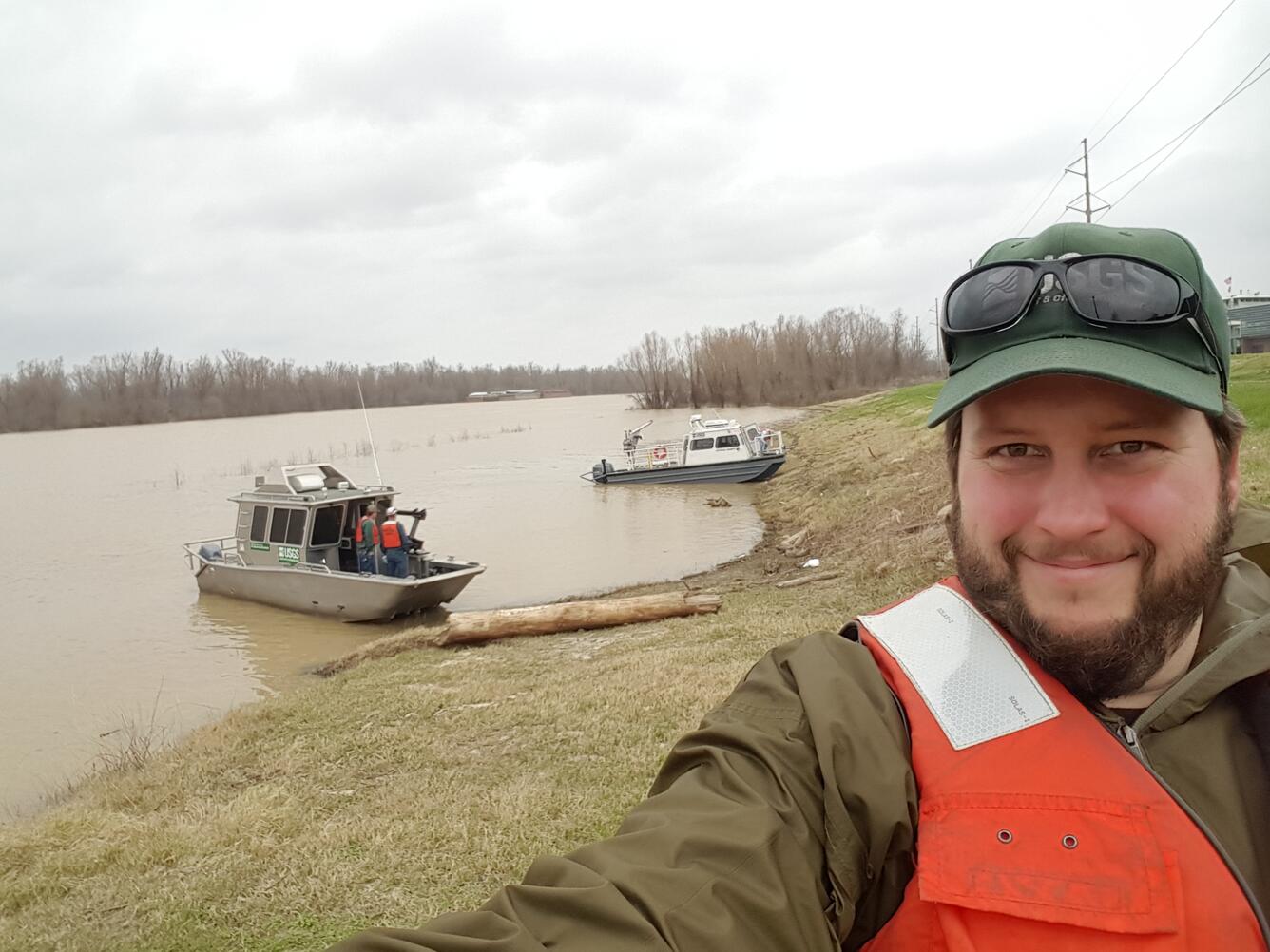 Hydrologic technician Dave Piatt takes a selfie in the field on a cloudy day. A river and boat can be seen in the background.