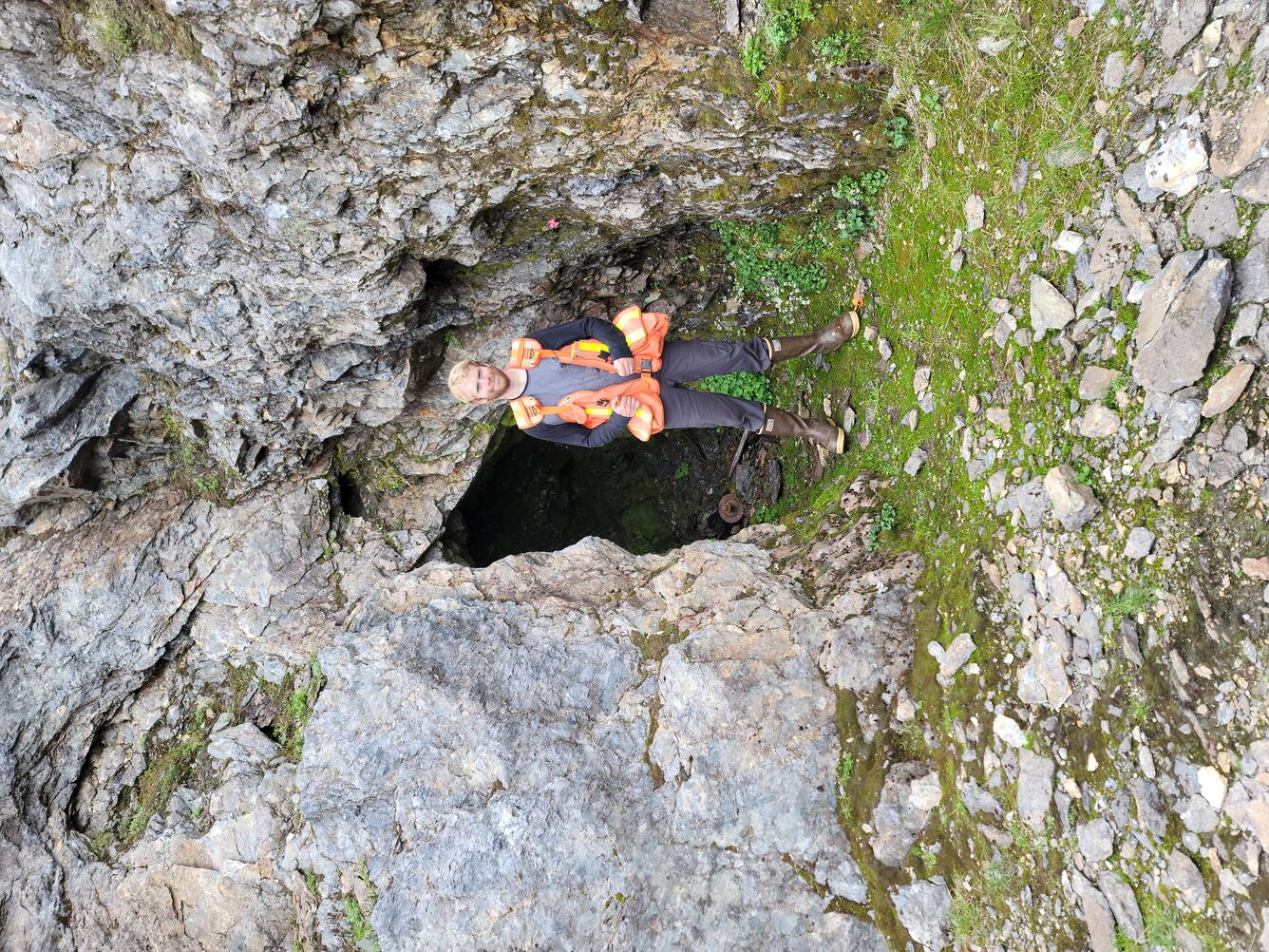 Geologist stands in front of an entrance to an underground mine wearing an orange vest with pockets and rubber boots.