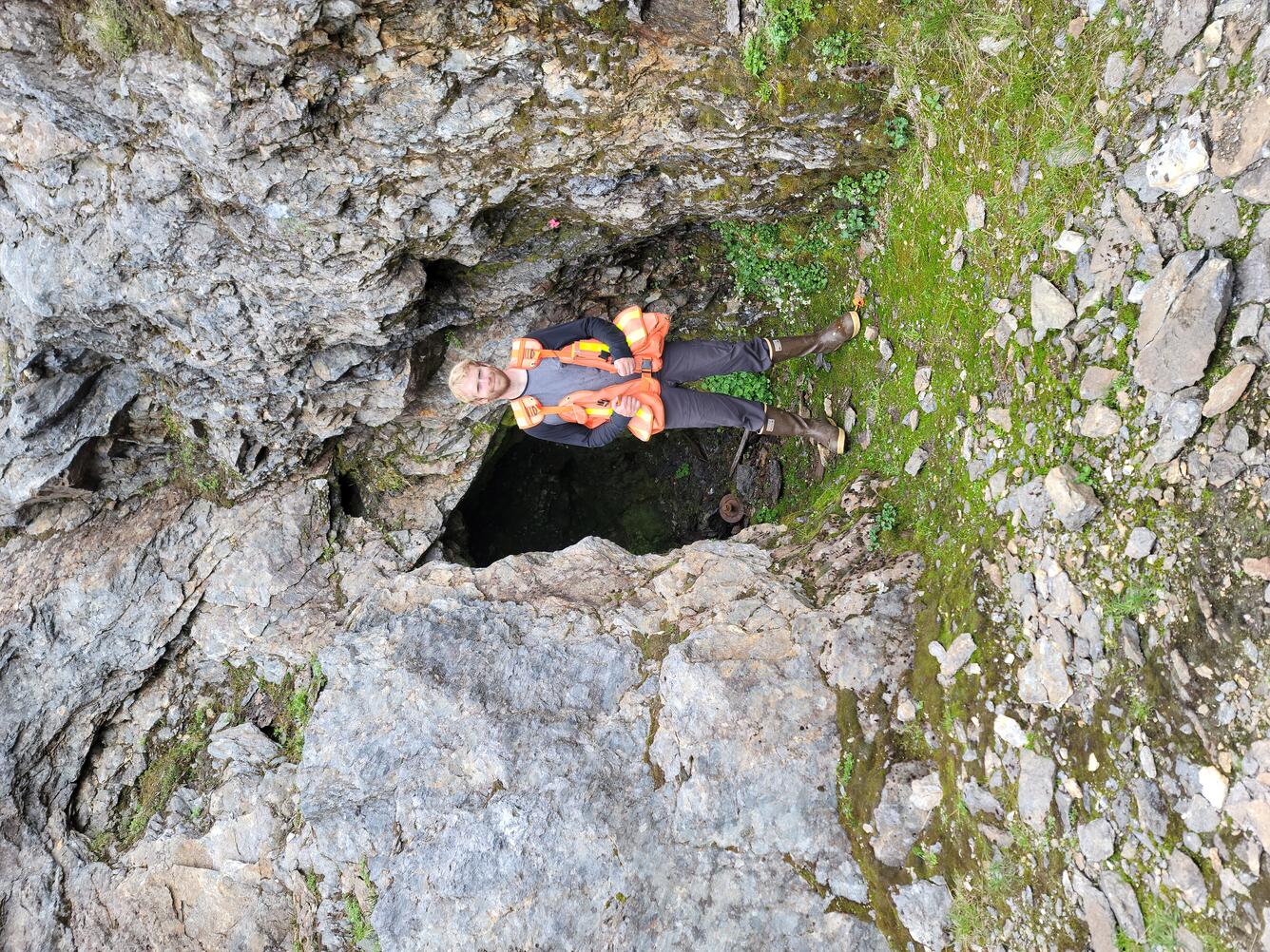 Geologist stands in front of an entrance to an underground mine wearing an orange vest with pockets and rubber boots. 