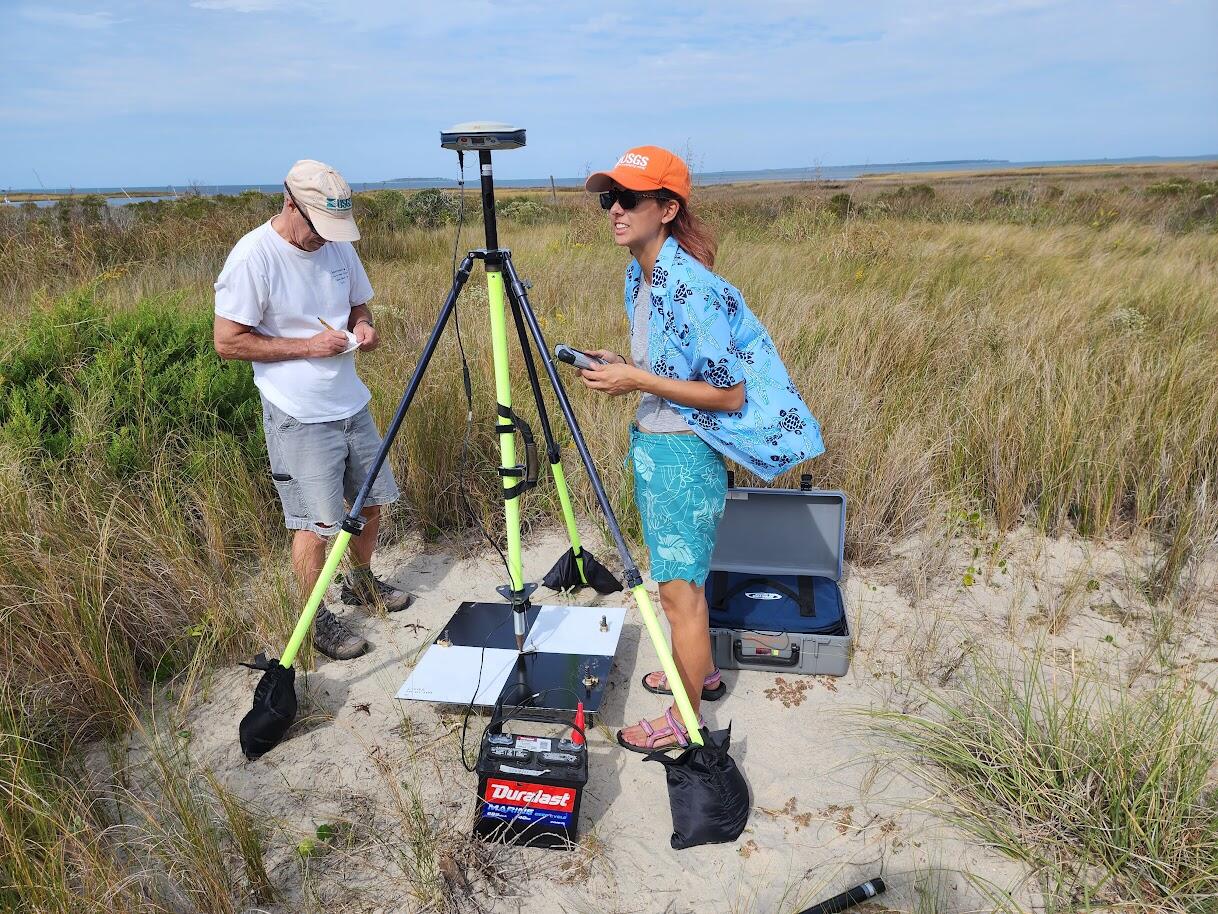 Two people on sandy beach surrounded by beach grass, next to a global navigation satellite system (GNSS) rover