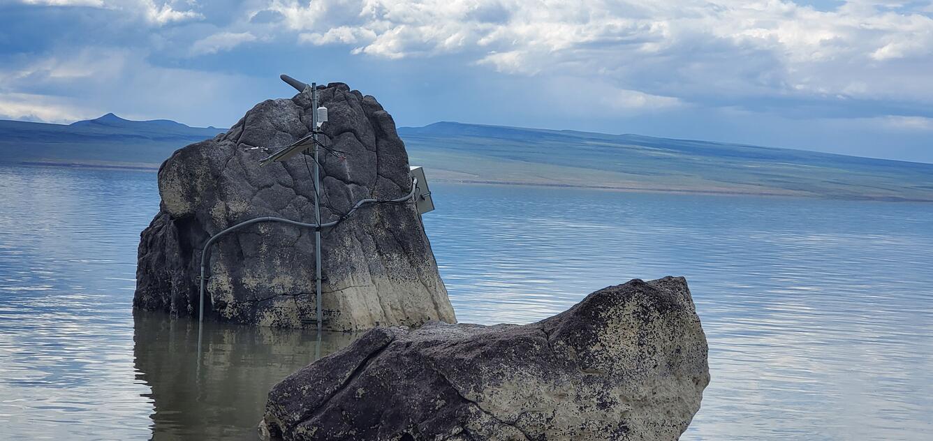 side view of grey DCP box , cables and telemetry antennas mounted to a large boulder in shallow blue water.