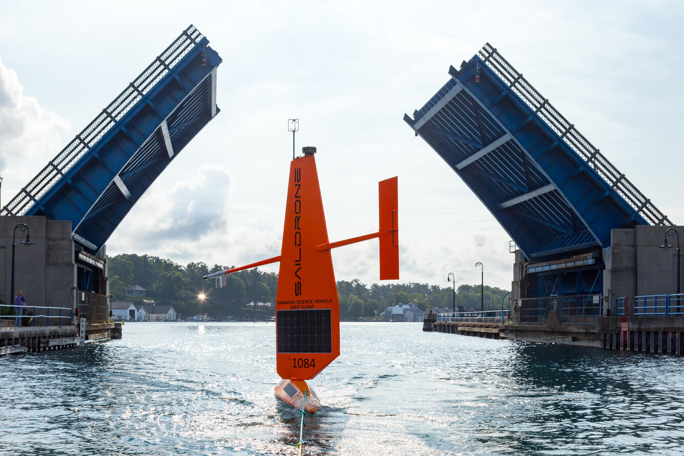 A saildrone, an orange autonomous sailing vessel, passes through an open drawbridge