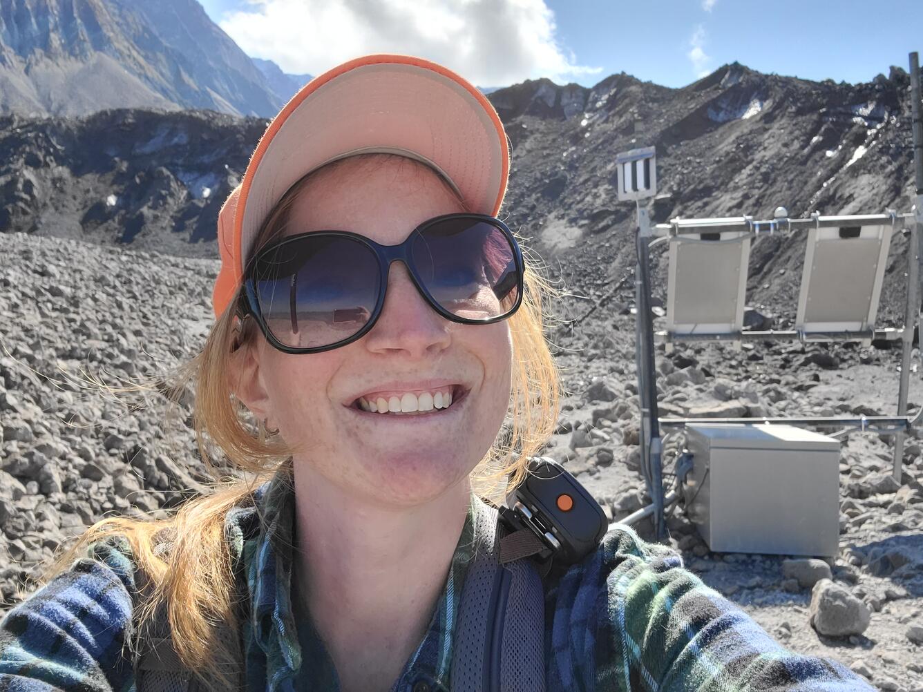 A young woman in dark sunglasses and an orange hat crouches next to a scientific sensor on a rocky slope. She has blonde hair and fair skin.