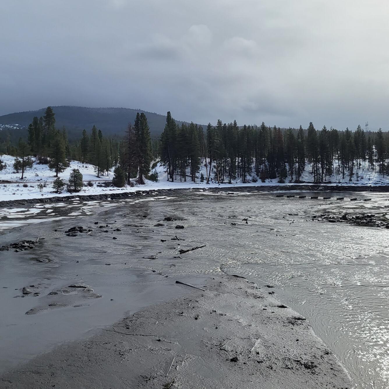 Klamath River flows through snow covered hills with sparse pine trees. Cloudy cold day