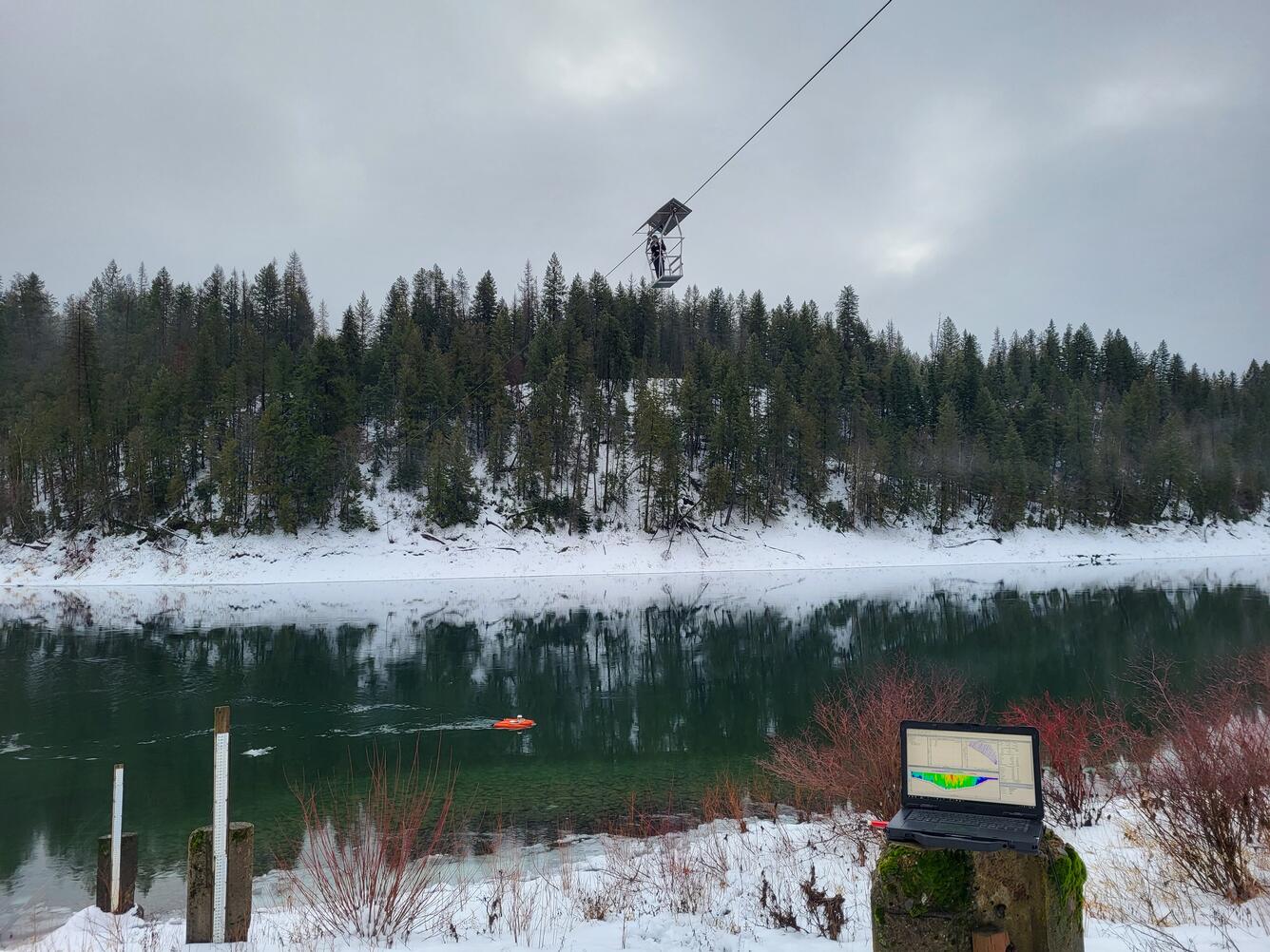 A technician is suspended above a river by cableway. In the foreground, scientific equipment lays on the snow.