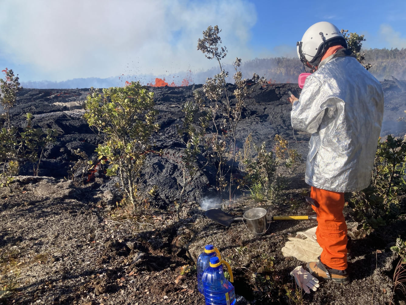 A scientist in a long silver heat-resistant coat, gas mask, and helicopter helmet inspects a just-quenched sample of lava