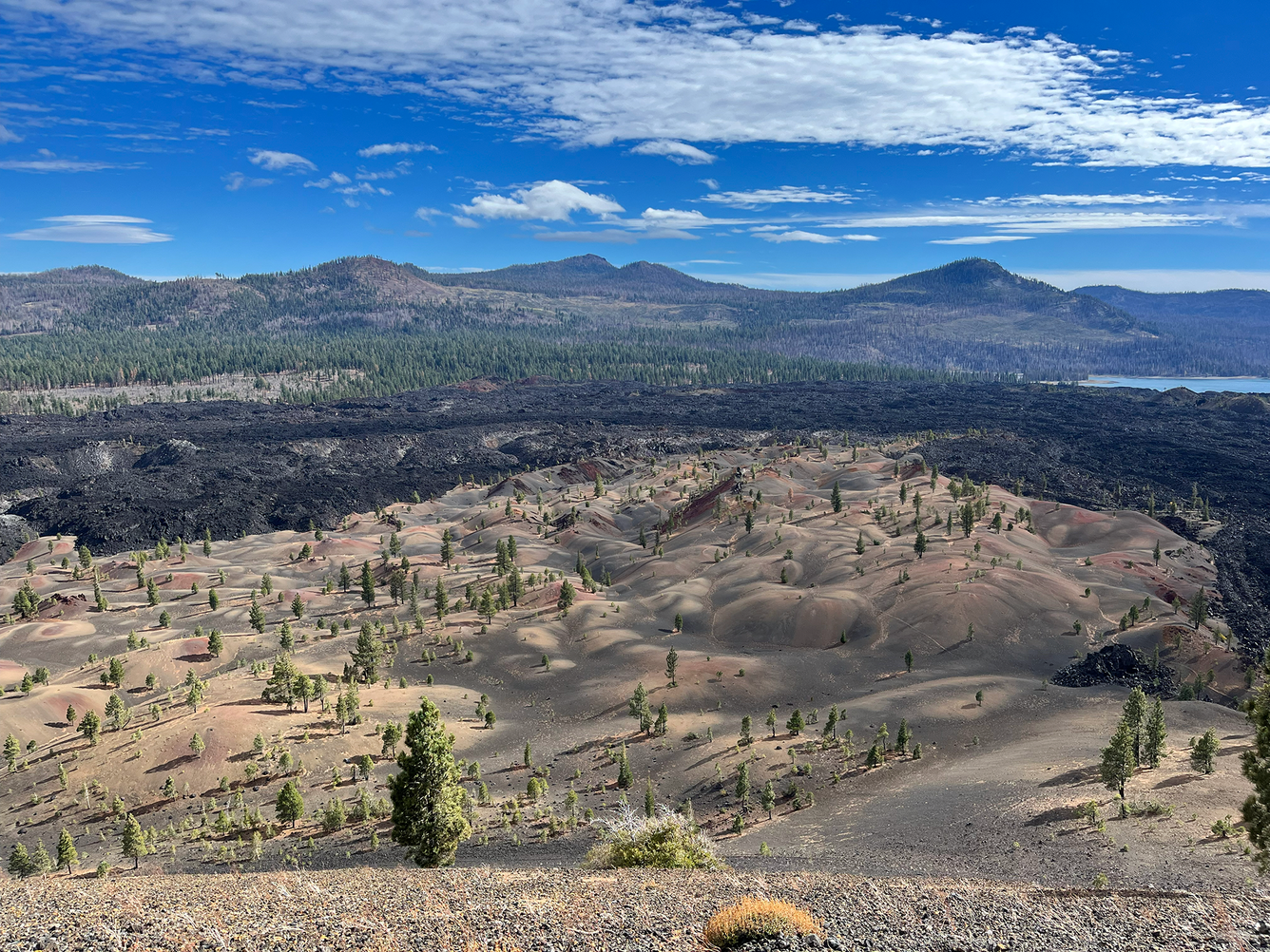 Taken from a high vantage point, this photo shows a landscape of rolling, barren hills stained in reds, tans, and blacks merging into a field of jagged black lava flows. In the distance, mountains rise above a pine-forest and bright blue lake.
