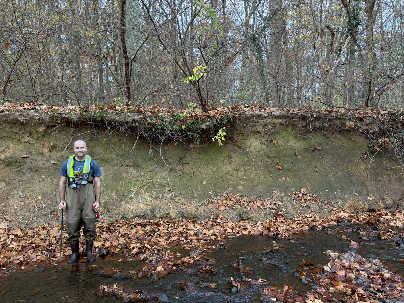 A USGS hydrologic technician stands in a creek with heavily eroded banks.