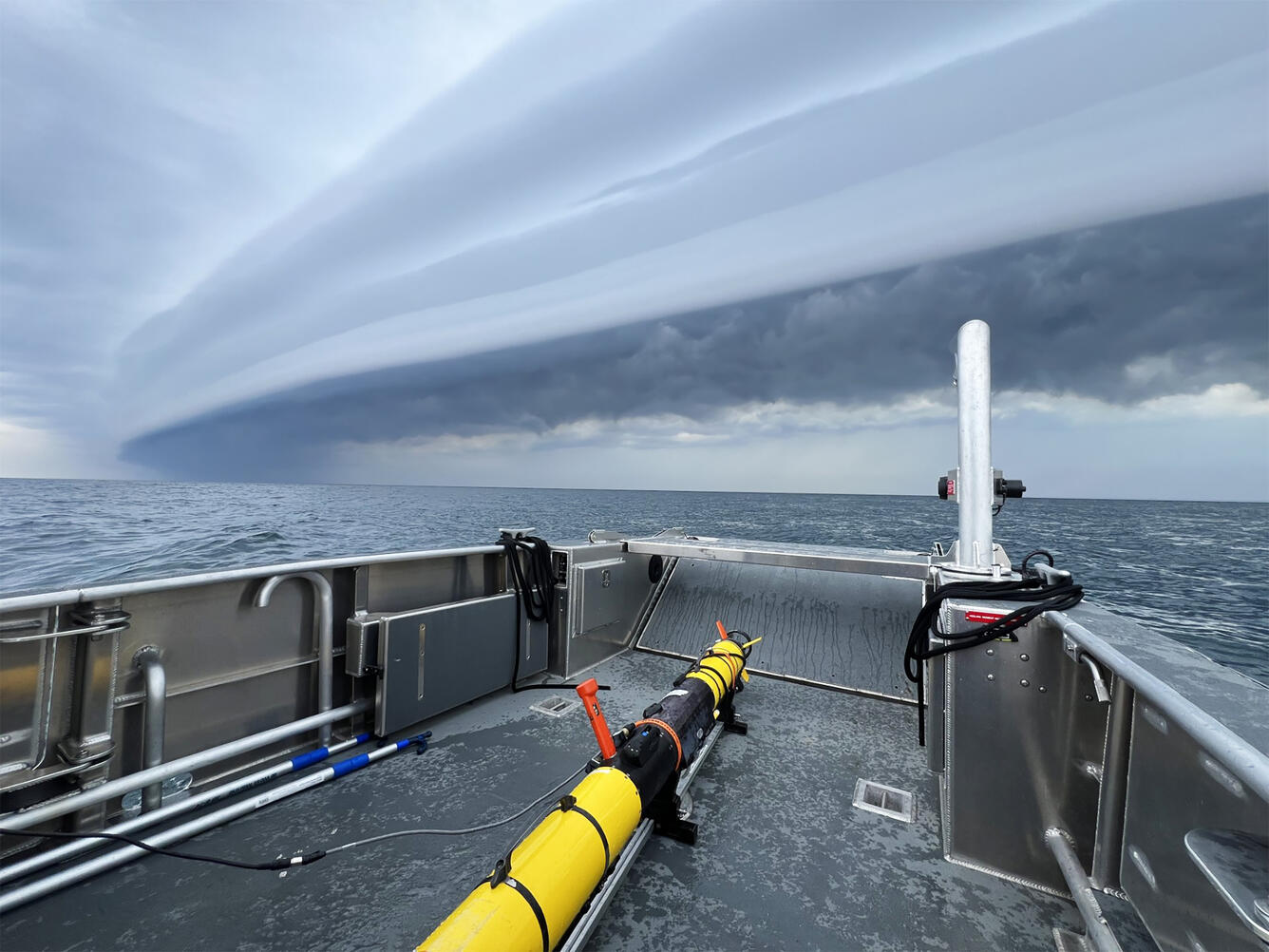 A long yellow autonomous vehicle sits on the deck of a research vessel on a stormy day.