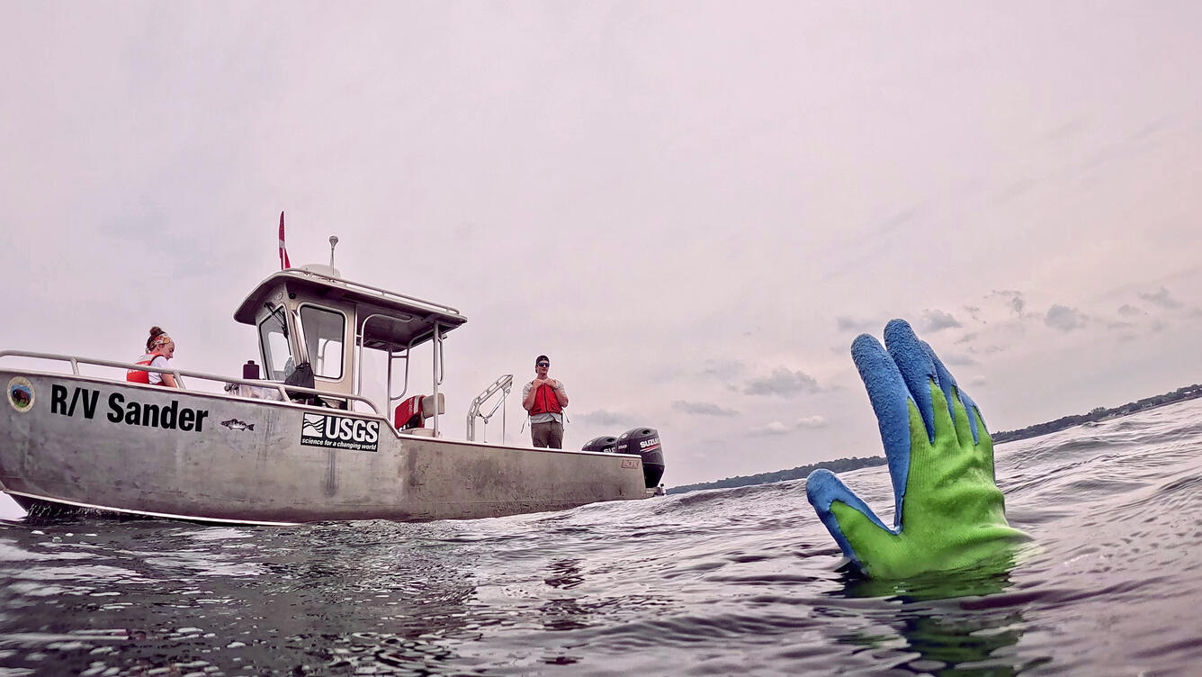 The gloved hand of a diver emerges from a lake and signals researchers on a small vessel, R/V Sander, in the background