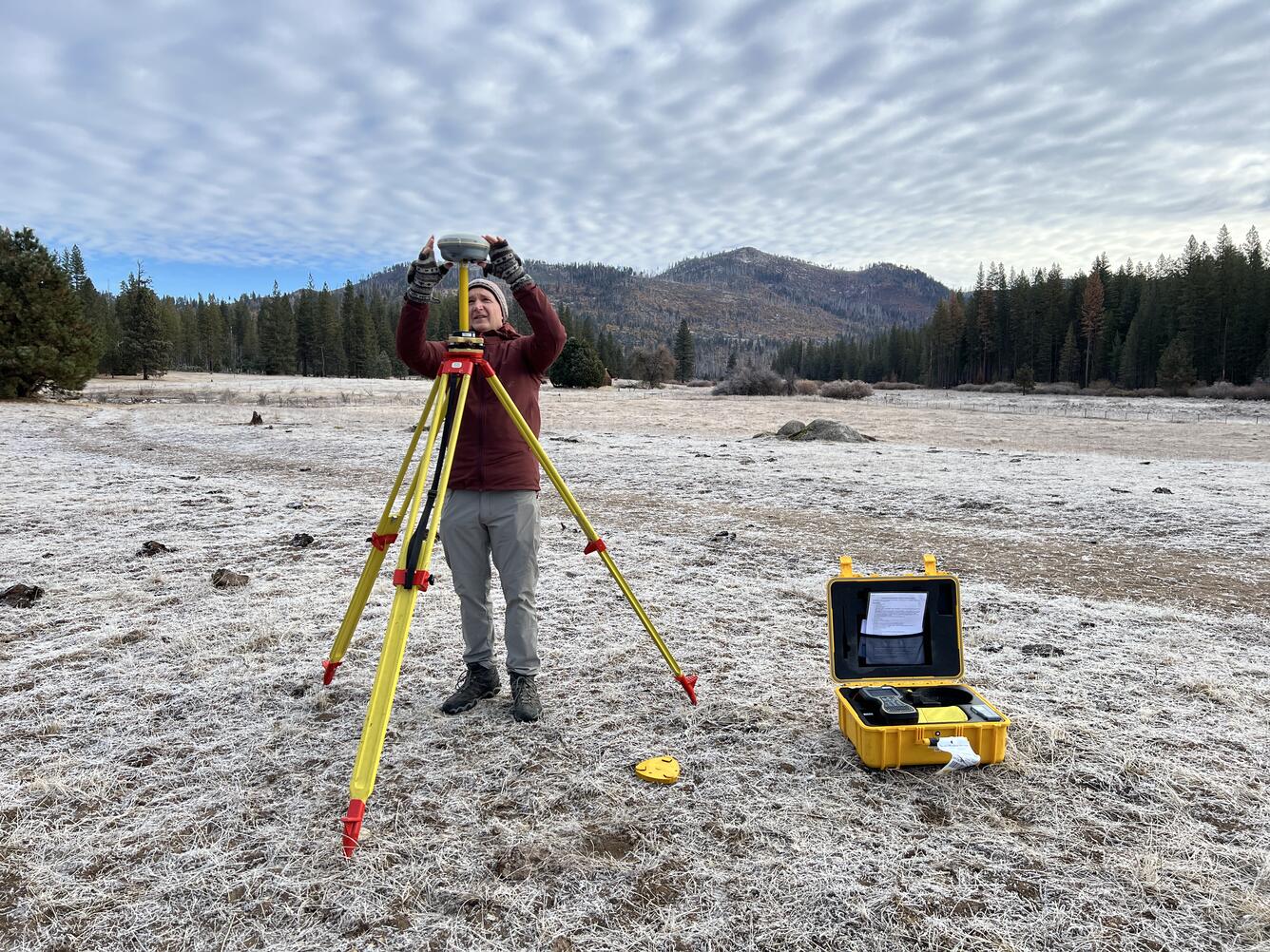 Setting up Global Navigation Satellite System base station on a tripod in a forest clearing with mountains in the background