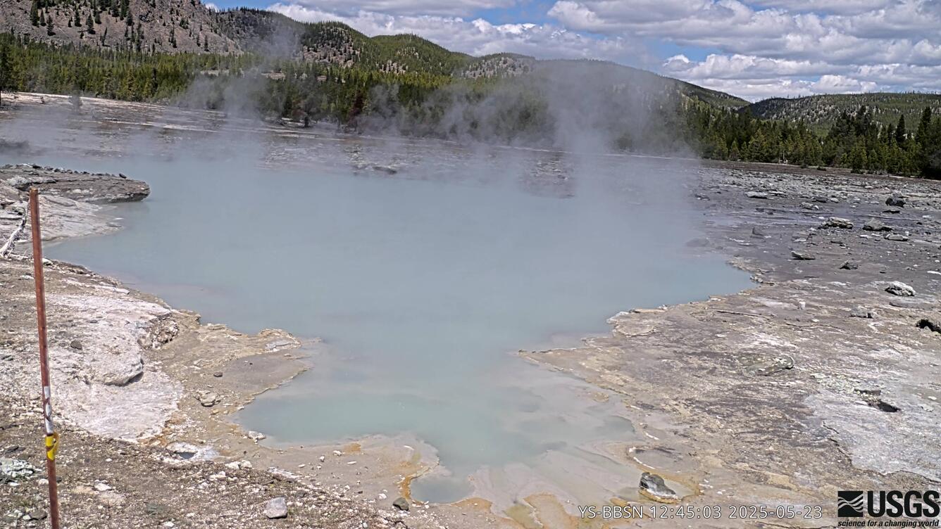 Webcam view of a steaming and murky pool surrounded by barren ground with rocky cliffs in the distance.