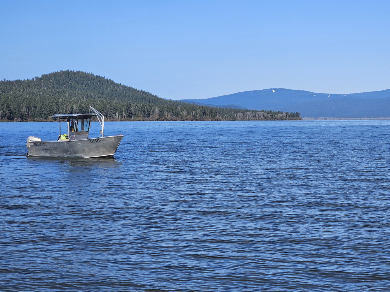 aluminum boat with a black USGS logo on vast blue lake waters. green forested hill in the background