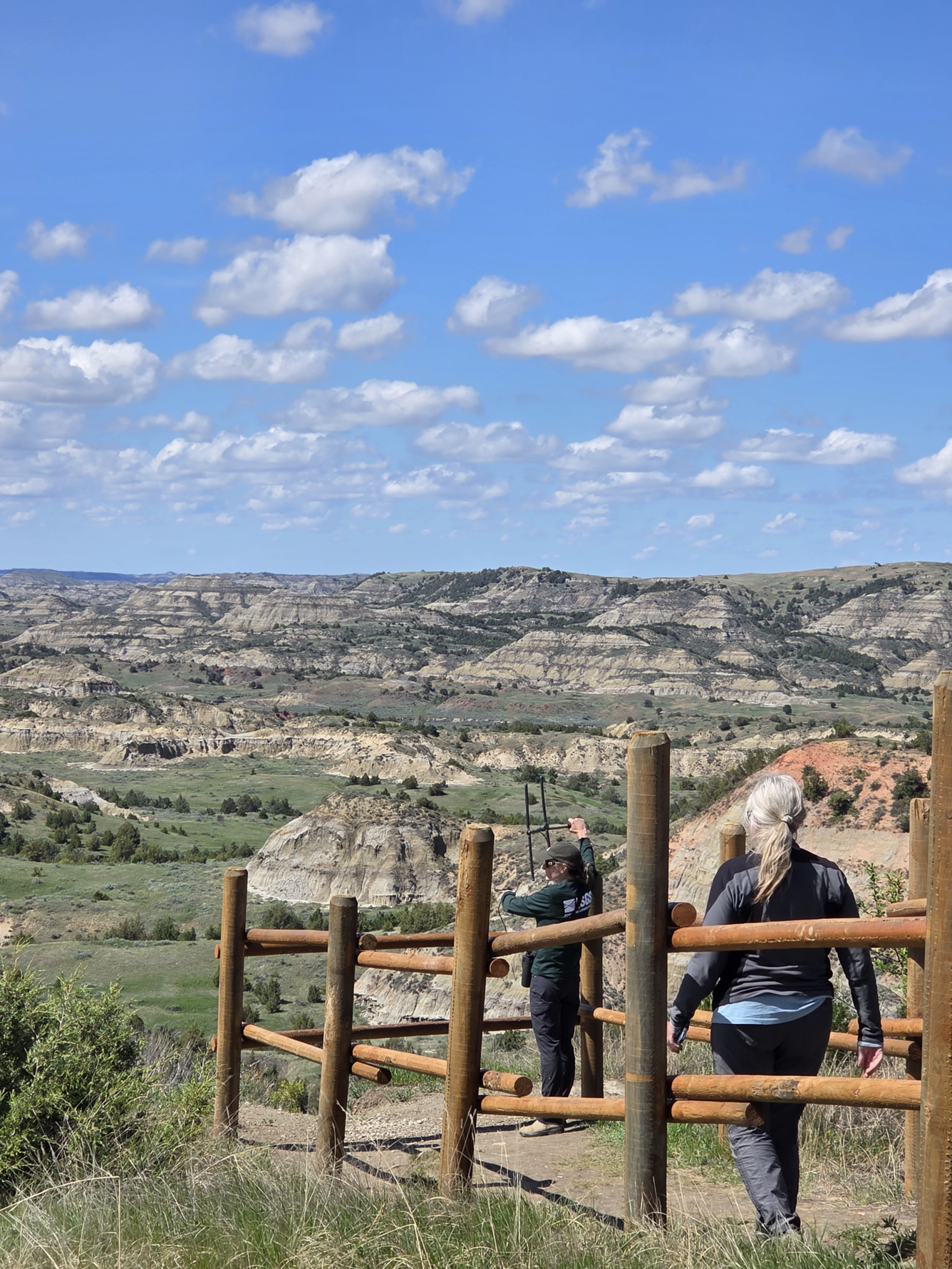 two people stand behind a fence, one holding up a telemetry device, mountains and sky in the background