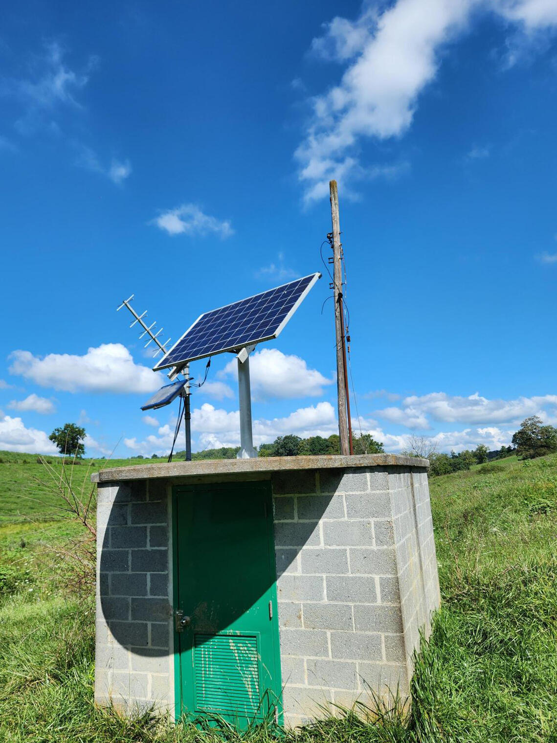 A small concrete block wellhouse with two solar panels and a satellite antenna on the roof.