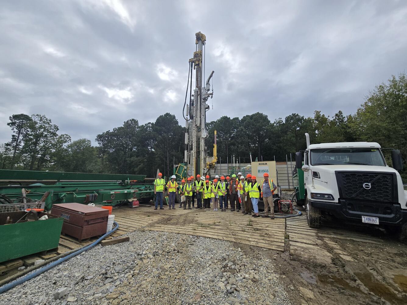 A tour group in safety equipment pose for a picture in front of a USGS drilling rig