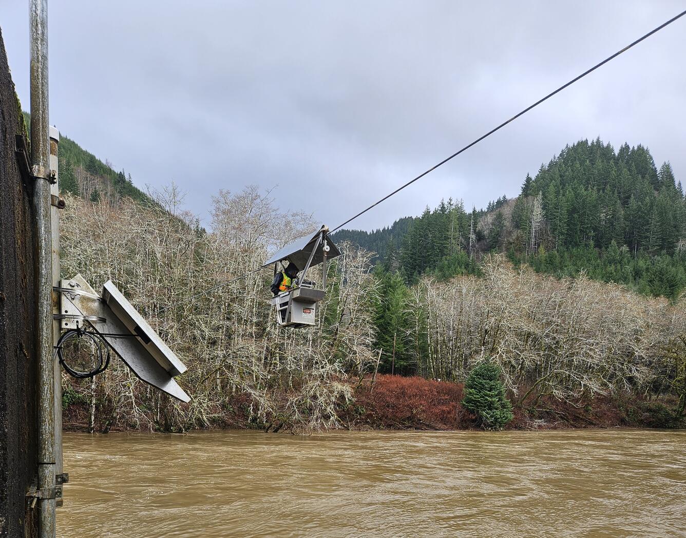 A man suspended above murky rushing river waters measures flow from an aluminum cable car