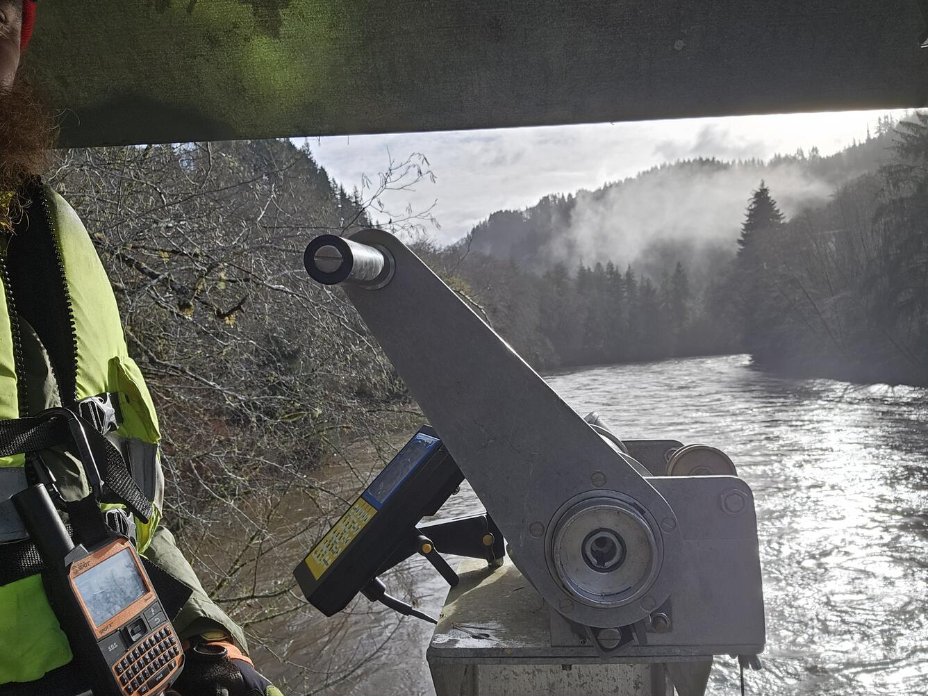 A man suspended above the river operates an aluminum crane used to lower measurement equipment into the water