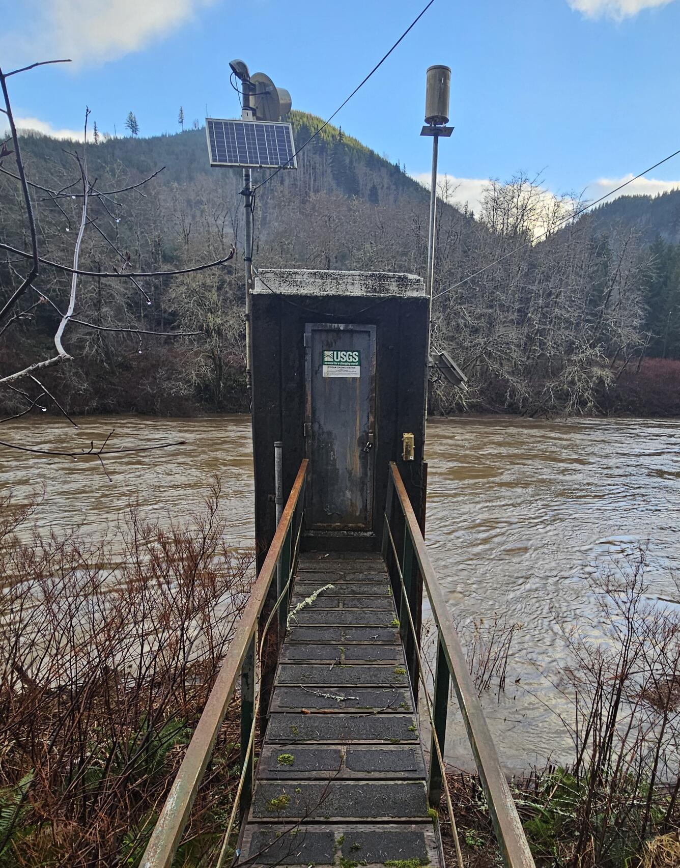 Square concrete structure built alongside the riverbank has solar power & antennas on top and wooden walkway leading to door