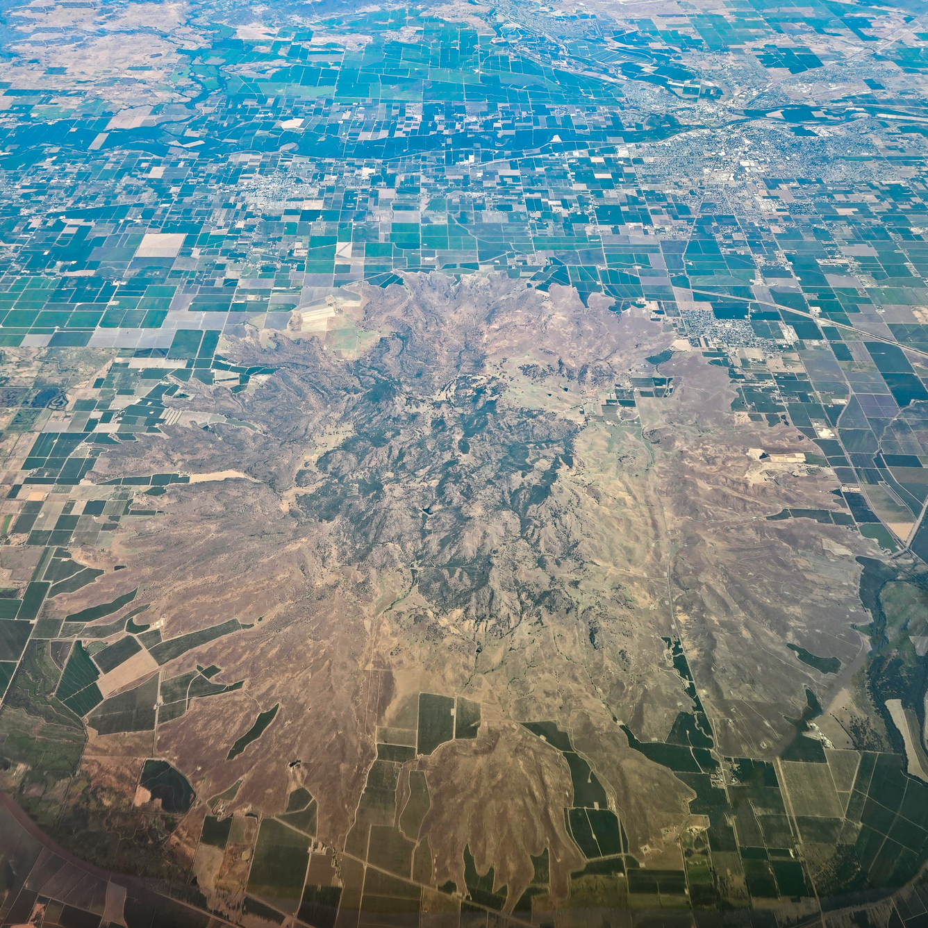 A photo taken from an airplane at high altitude shows a cluster of craggy, brown and gray volcanic peaks surrounded by a circular apron of sloping, vegetated debris. Surrounded the volcano are green and brown agricultural fields laid out in a grid pattern.