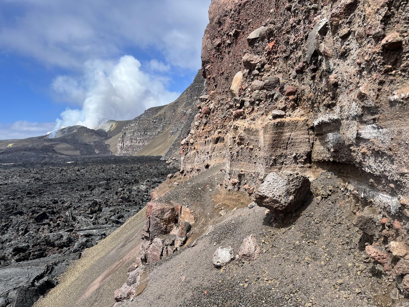 Color photograph of explosive volcanic deposits in wall of crater with degassing volcanic vents in the background